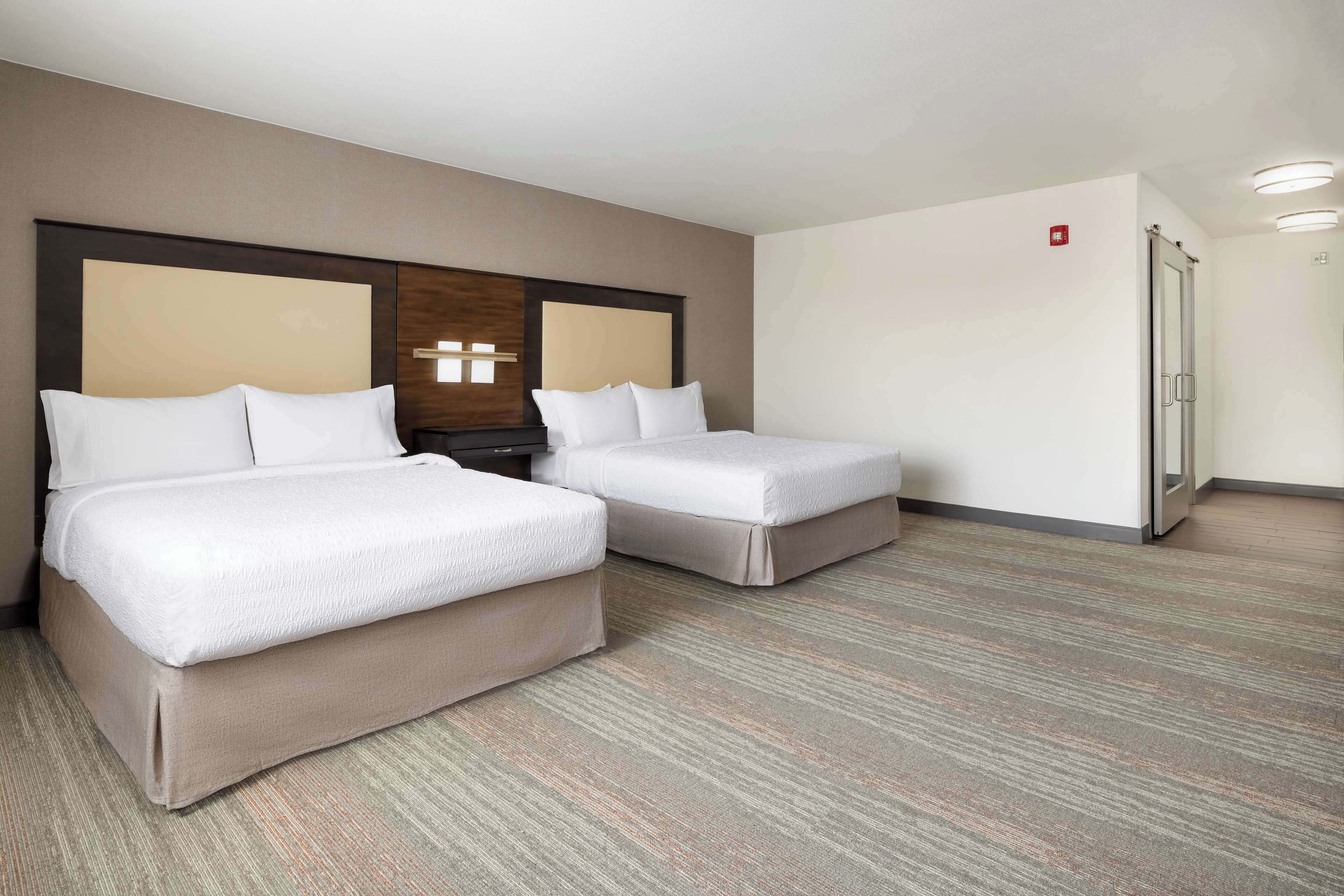 A hotel room with two beds, a wooden headboard, a nightstand, a refrigerator, and a carpeted floor.