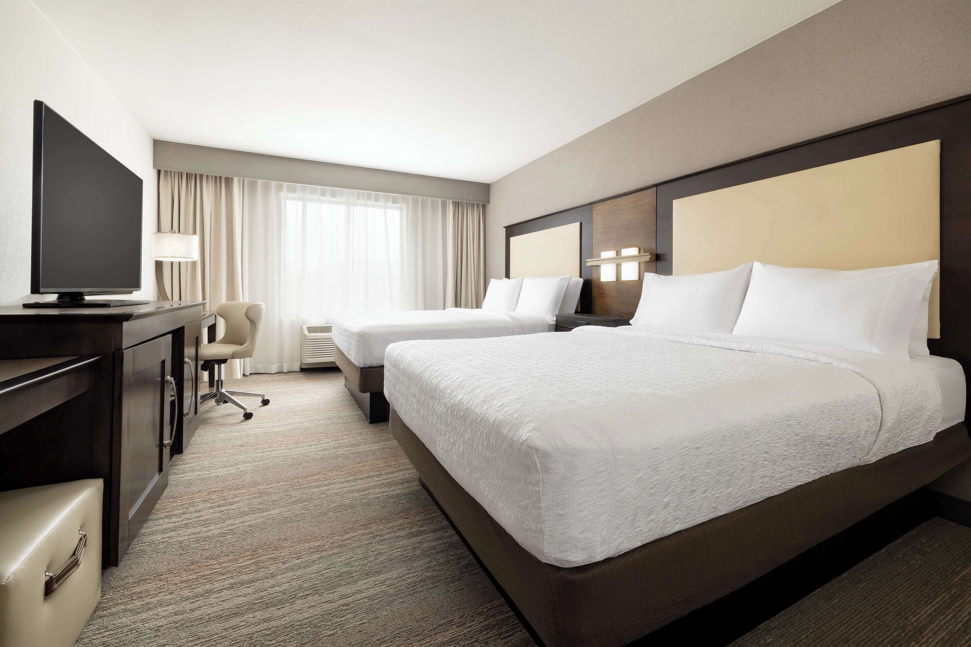 A hotel room with two beds, a desk with a flat-screen TV, a chair, and a lamp.