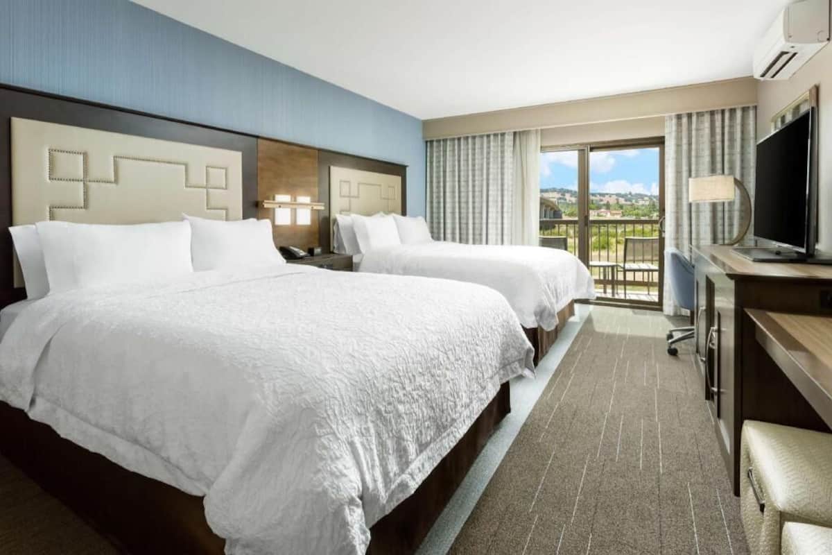 A hotel room with two beds, a desk, a TV, and a balcony with a view.