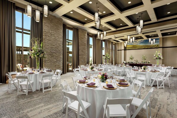 A banquet hall with round tables set for dining, white chairs, and large windows allowing natural light.