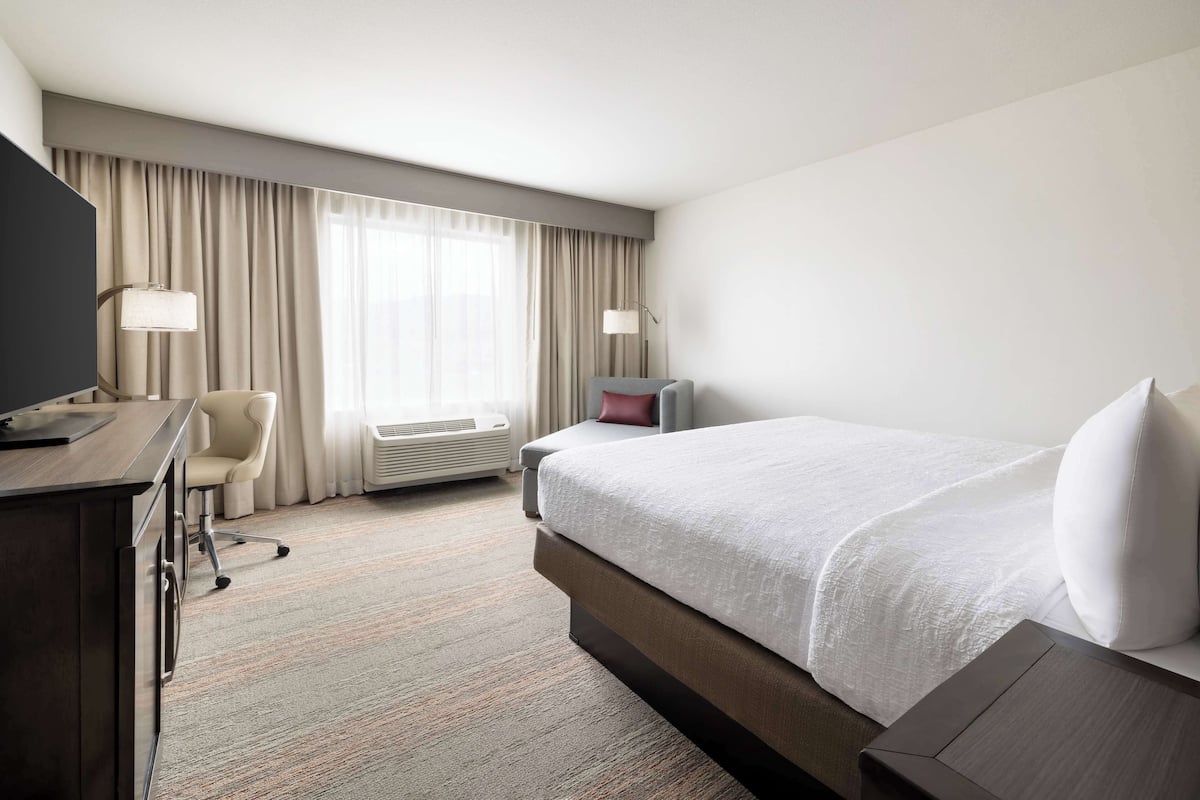 A hotel room with a bed, desk, chair, and a large window with curtains.