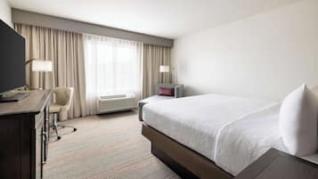 Una habitación de hotel con cama, escritorio, silla y un ventanal con cortinas.