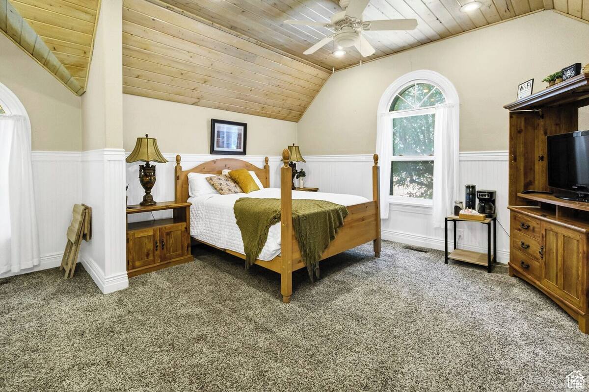 A bedroom with a wooden four-poster bed, a ceiling fan, a small table with a lamp, a TV stand, and a window with a view of trees.
