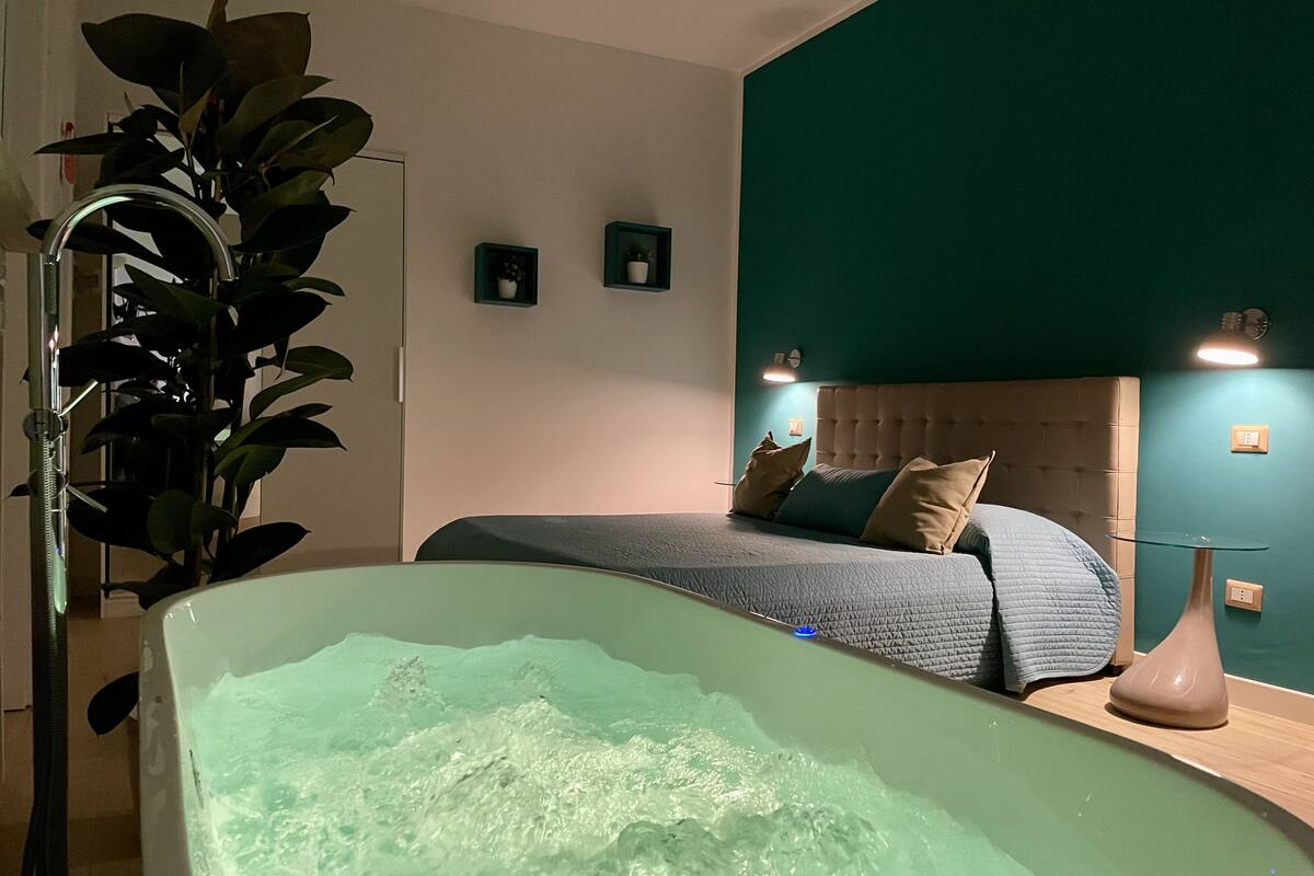 A modern bedroom with a large bathtub, a bed with a tufted headboard, and a potted plant.