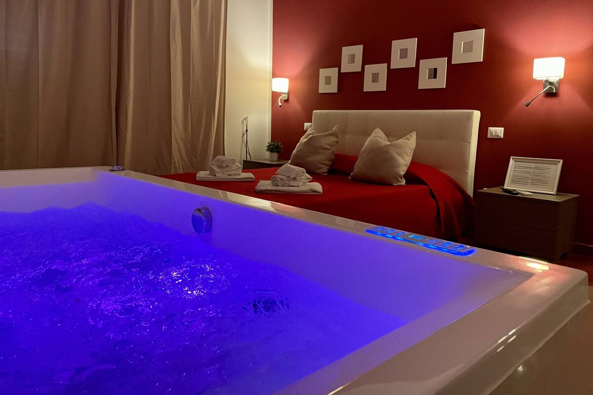 A hotel room with a large bathtub, a red bed with white pillows, and wall-mounted lights.