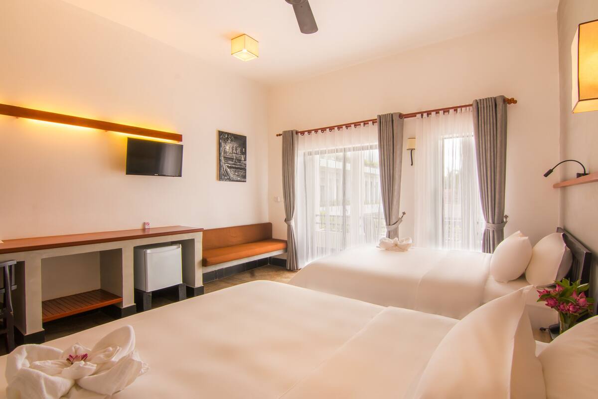 A hotel room with two beds, a desk, a TV, and a window with curtains.