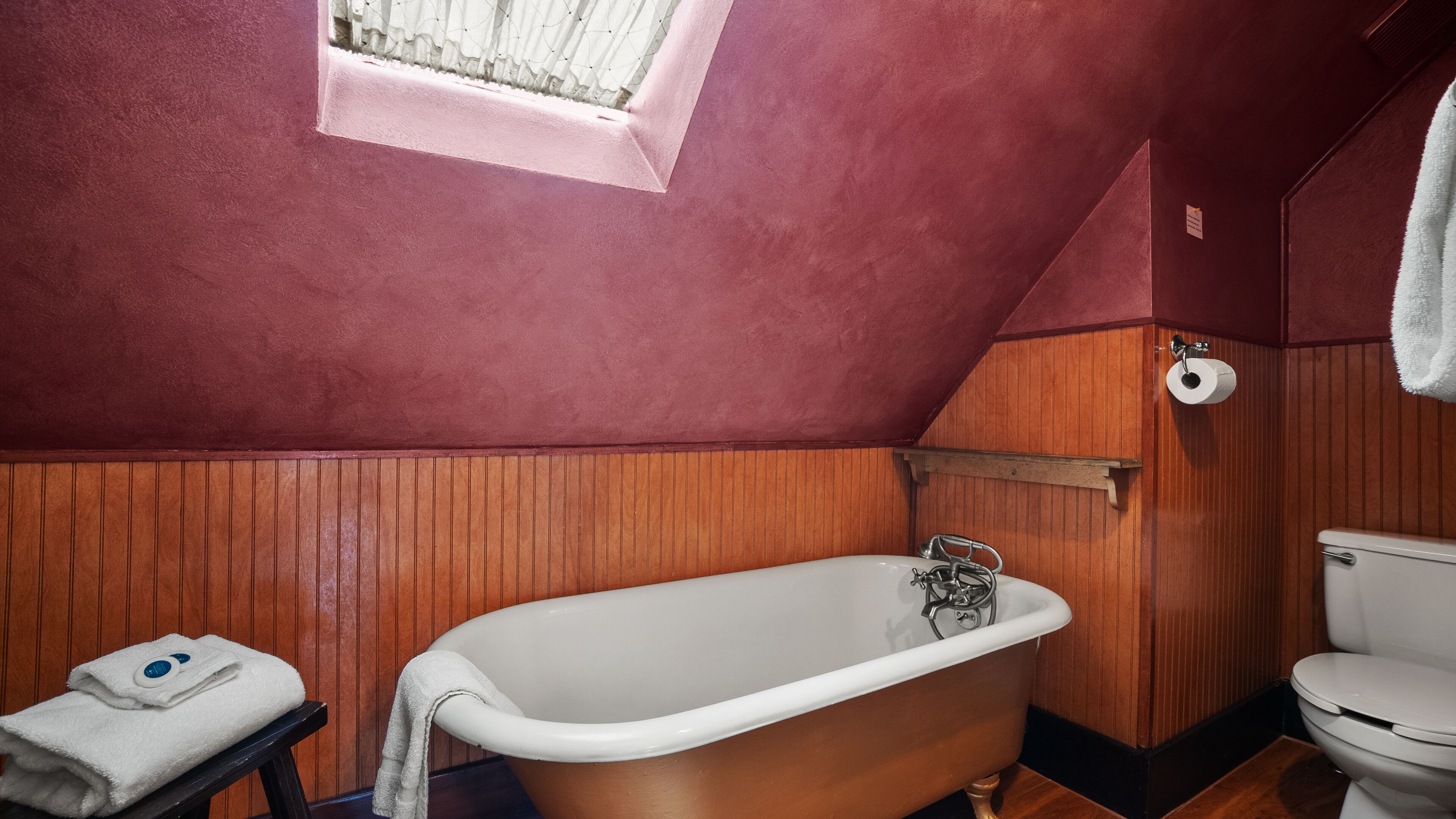 A bathroom with a clawfoot bathtub, wooden paneling, a toilet, and a window with a curtain.