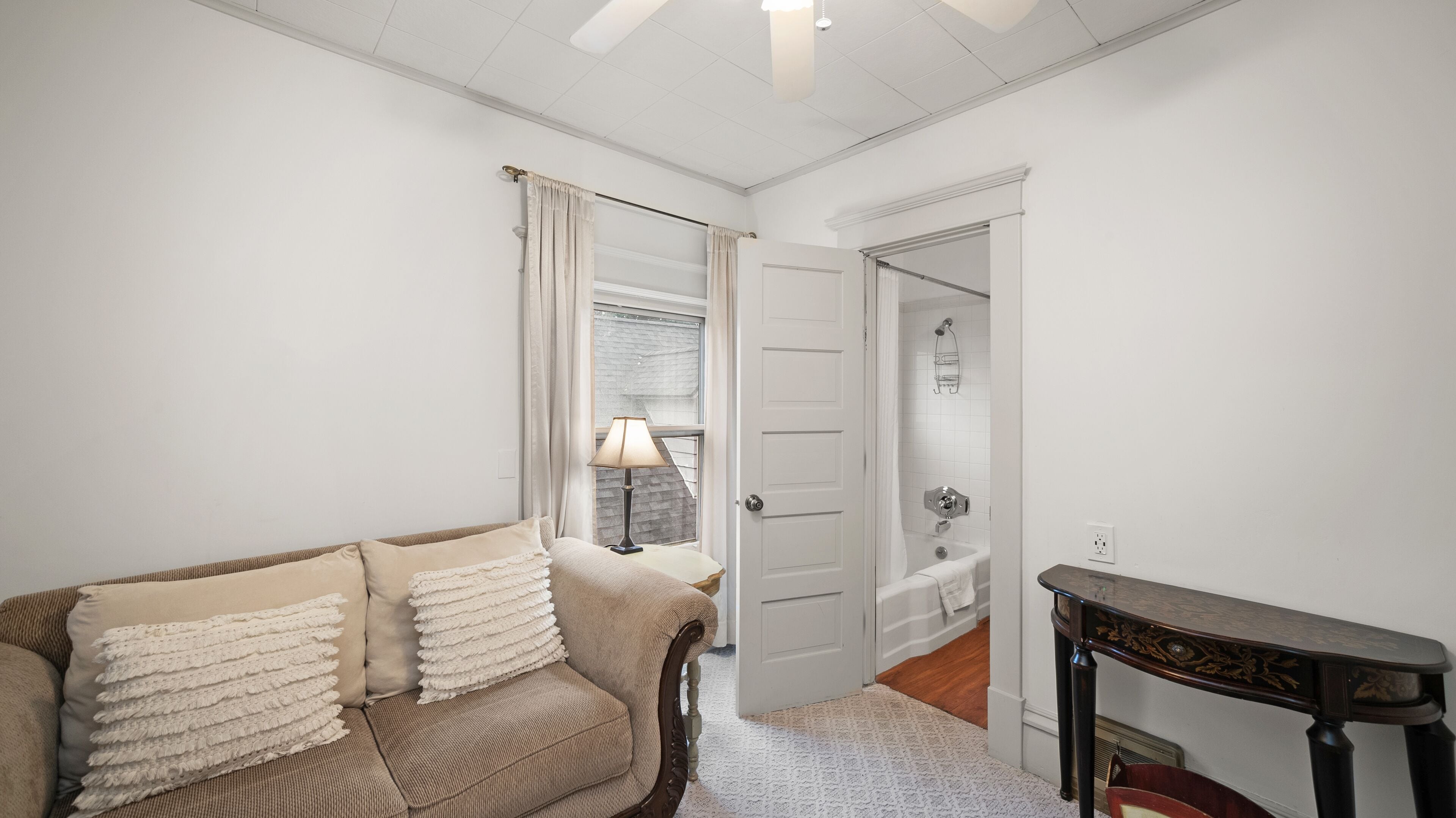 A room with a beige sofa, a lamp, a wooden table, and a door leading to a bathroom.