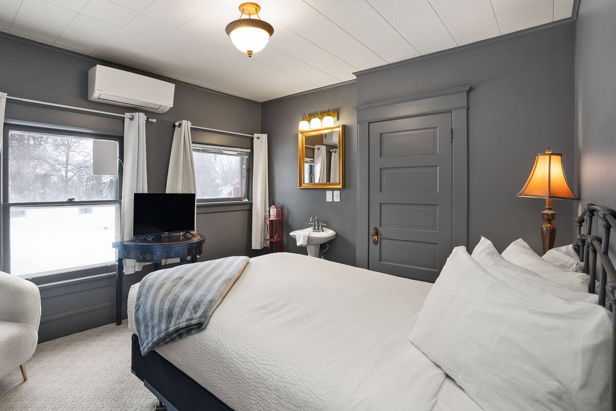 A hotel room with a bed, a chair, a desk with a TV, a window, a mirror, and a lamp.