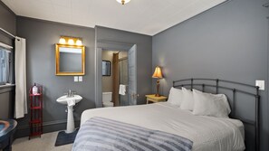 A bedroom with a bed, a bedside table, a lamp, a mirror, and a sink.