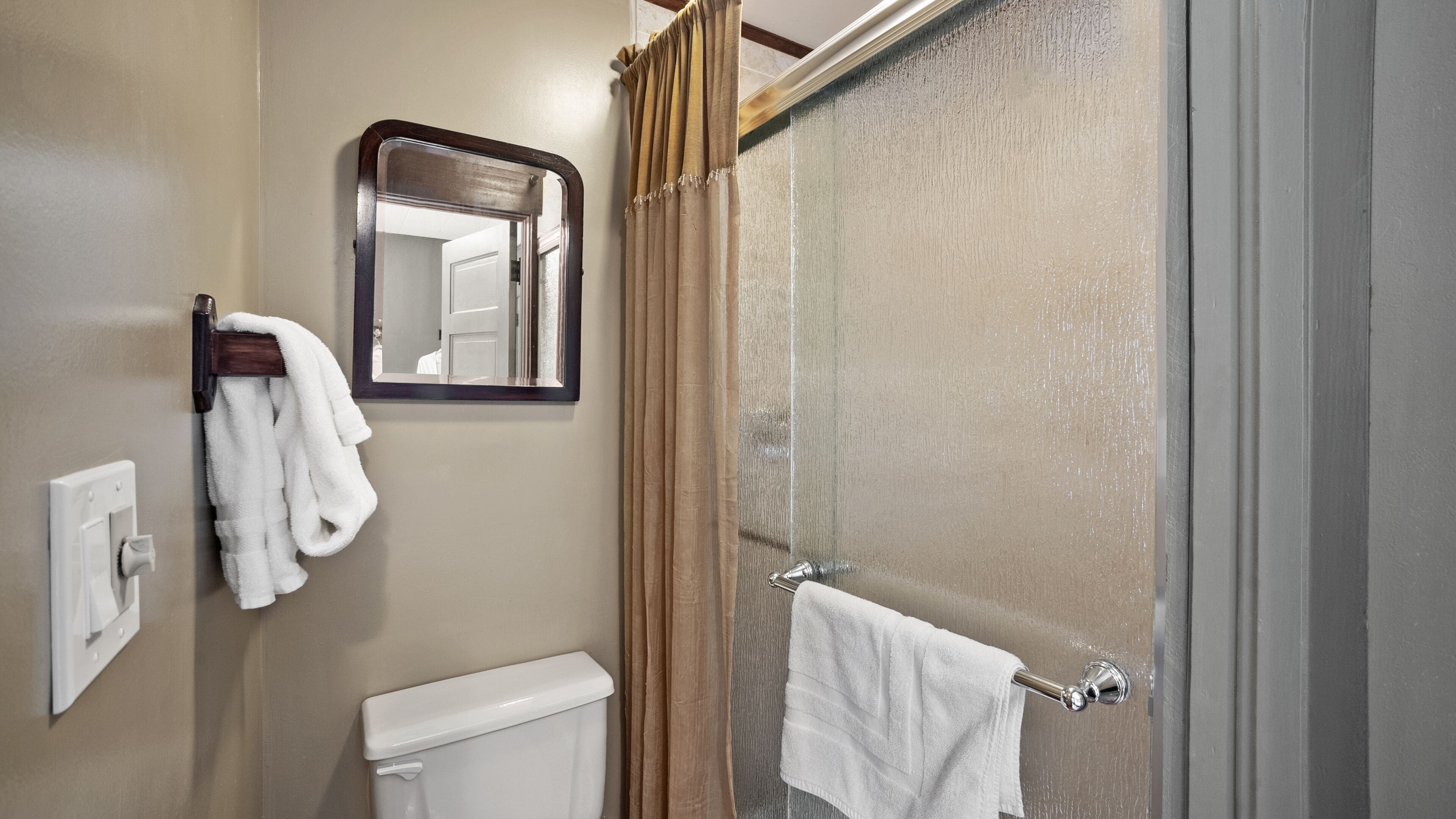 Un baño con inodoro, toallero con toalla blanca, espejo y ducha con puerta de cristal.