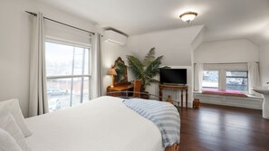 A hotel room with a large bed, a desk with a TV, a plant, and a window with a view of the street.