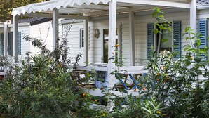 A white mobile home with blue shutters, a covered porch, and a garden area with various plants and flowers. - Camping Flumendosa (Pula)