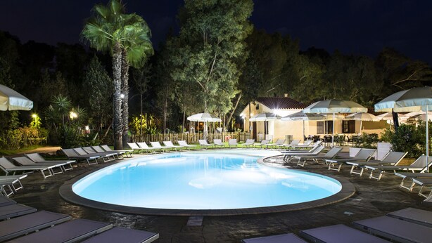 A nighttime pool area with lounge chairs and umbrellas, surrounded by trees and a building with a sign that reads "ALORIA". - Camping Flumendosa (Pula)