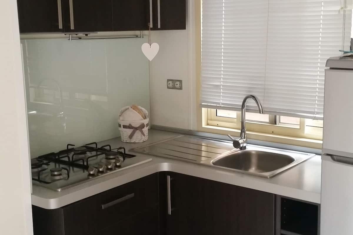 Een kleine keuken met donkere kastjes, een gasfornuis, een spoelbak en een raam met jaloezieën.
