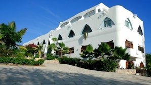 A modern building with a white facade, multiple arched windows, and a paved entrance area with tropical plants. - Watamu Palm Beach (Watamu)