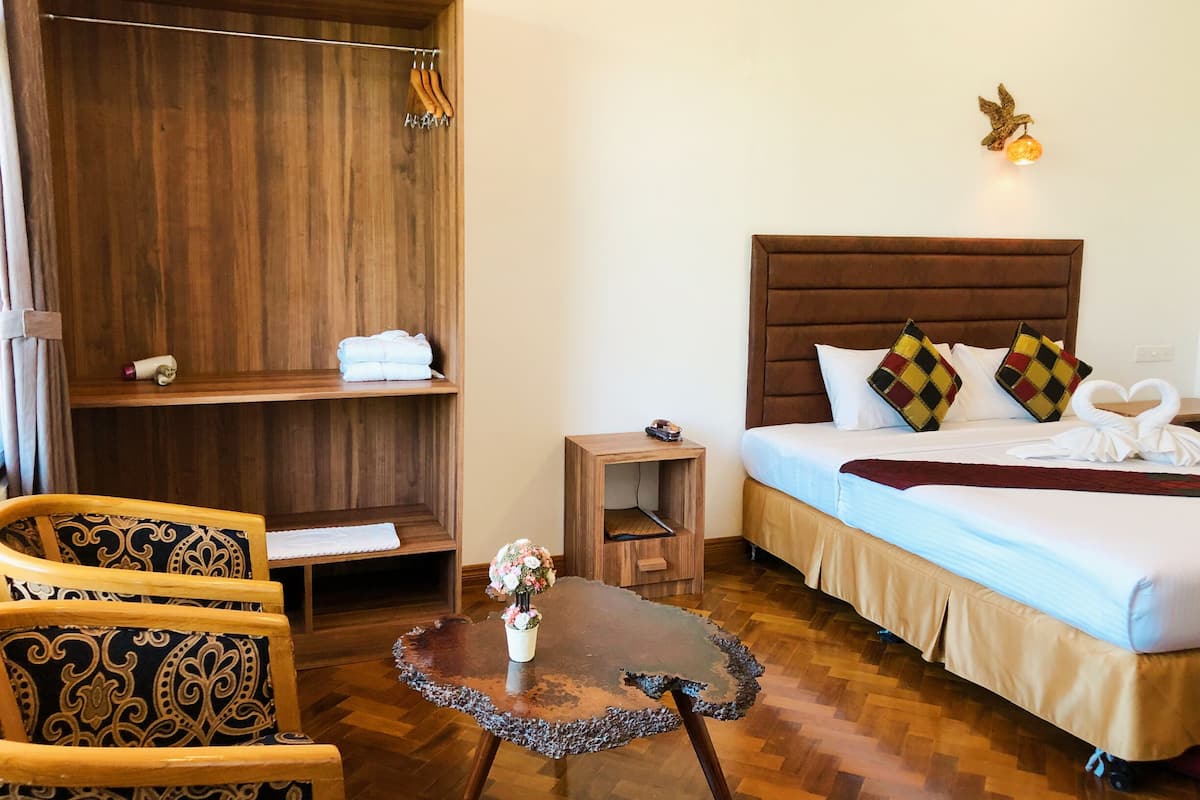 Une chambre d’hôtel avec une armoire en bois, un lit agrémenté d’oreillers à motifs, une petite table sur laquelle est posé un vase de fleurs et une chaise avec un coussin à motifs.