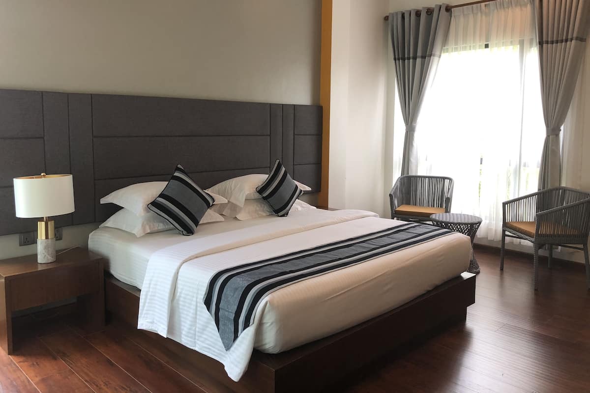 Un lit bien fait, recouvert d’une couverture à rayures, une lampe de chevet et une chaise près de la fenêtre, derrière des rideaux légers.