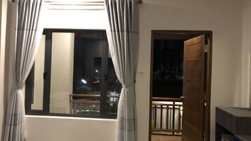 A room with a large window, curtains, a wooden door, and a view of a balcony.