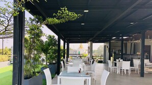 Un café al aire libre con mobiliario de mimbre blanco, techo cubierto y plantas en macetas.