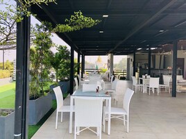 Un café al aire libre con mobiliario de mimbre blanco, techo cubierto y plantas en macetas.