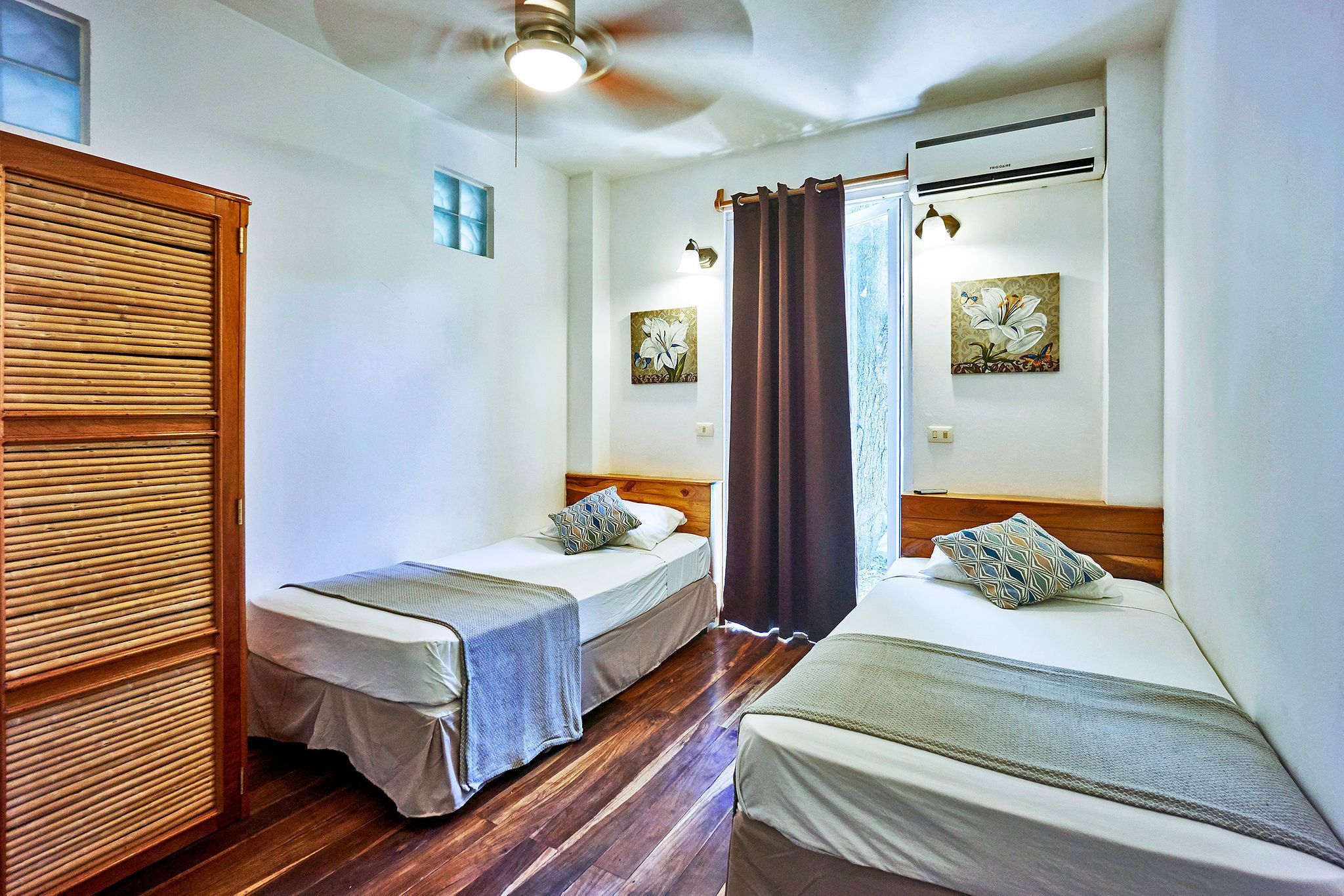 A room with two single beds, wooden flooring, a ceiling fan, and two wall-mounted air conditioning units.