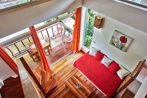 Una habitación con cama roja, suelo de madera, balcón con sillas y vistas al exterior con vegetación.