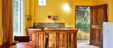 A kitchen with wooden cabinets, a dining table with chairs, a small window, and a view of greenery outside.