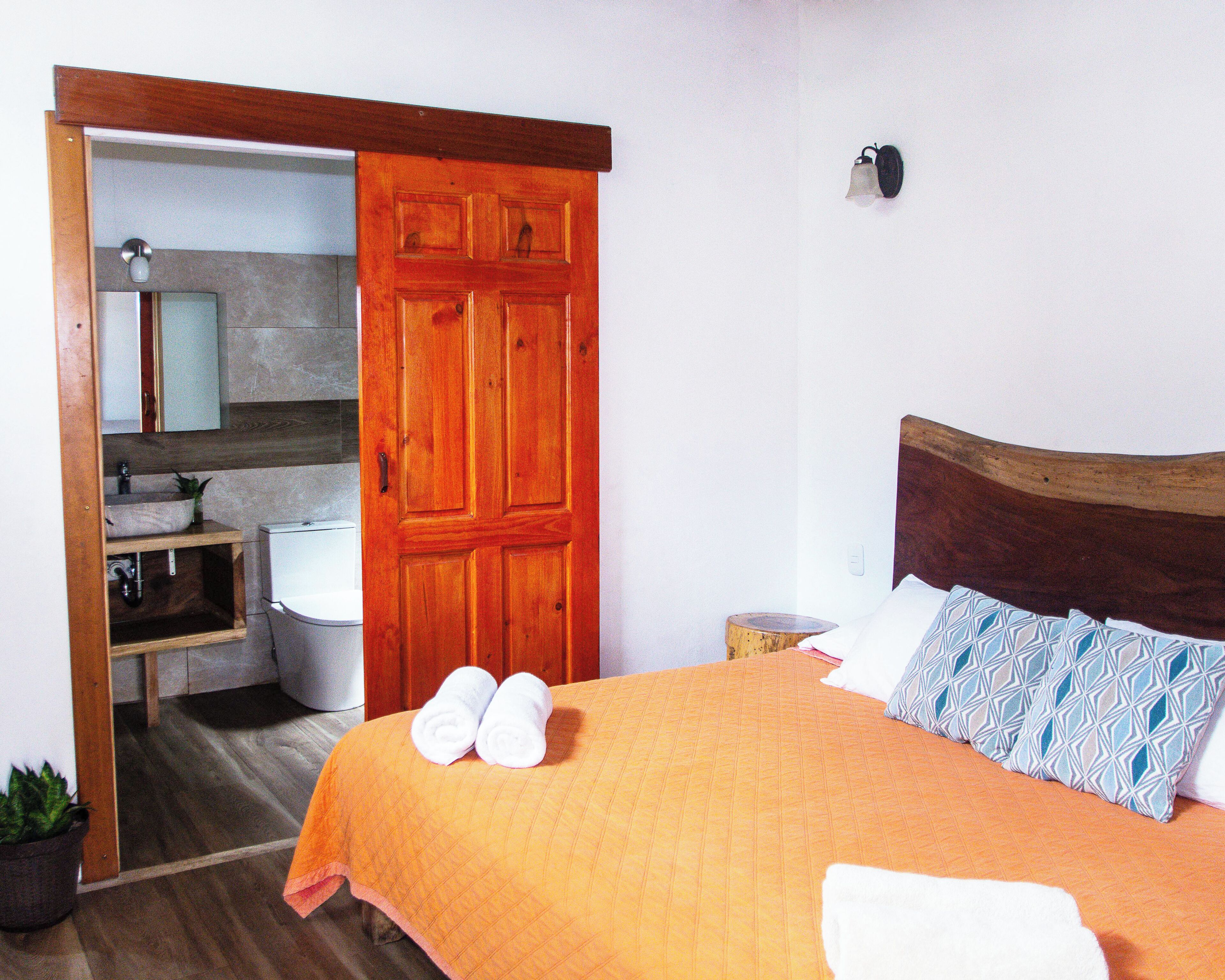 Un dormitorio con puerta de madera, cama con colcha naranja, dos toallas blancas, mesita de noche y una lámpara de pared.