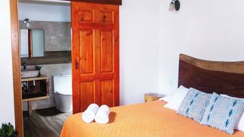 Un dormitorio con puerta de madera, cama con colcha naranja, dos toallas blancas, mesita de noche y una lámpara de pared.