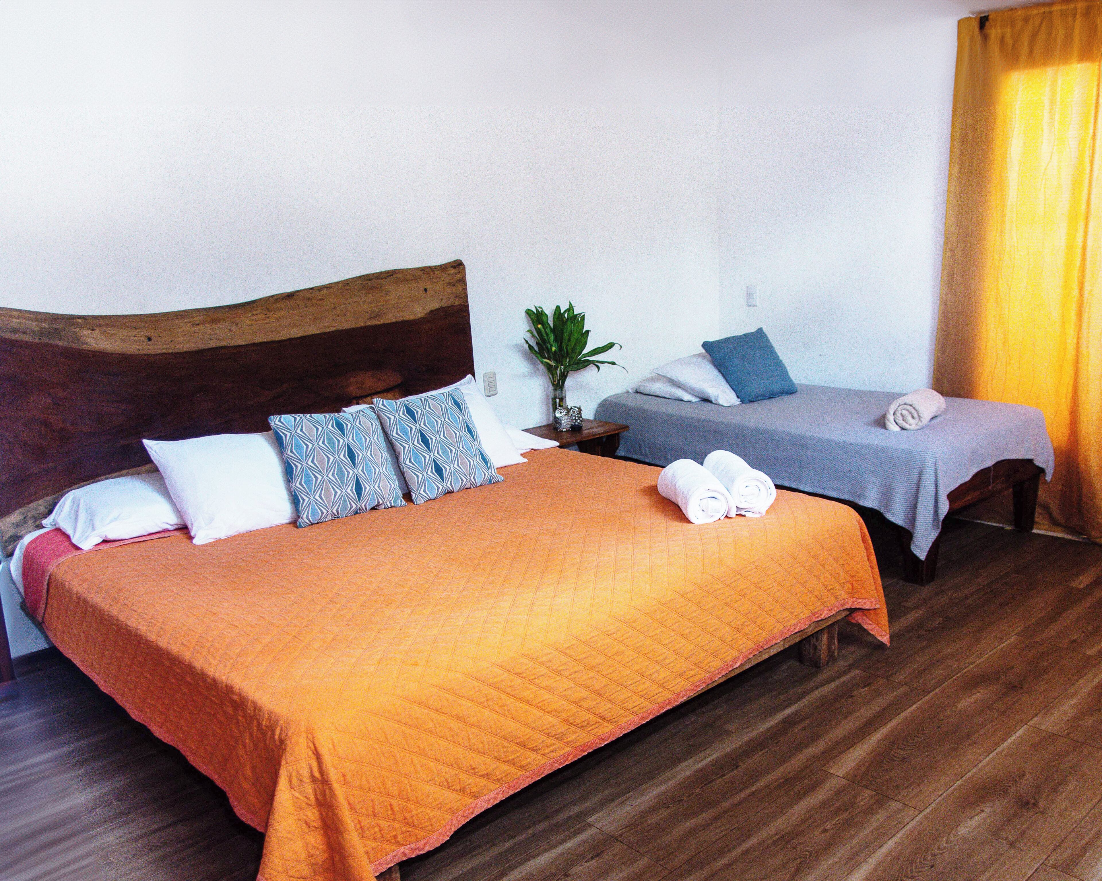 Une chambre d’hôtel avec un grand lit, un plus petit lit, un plancher en bois, une table de chevet avec une plante et une fenêtre avec des rideaux jaunes.