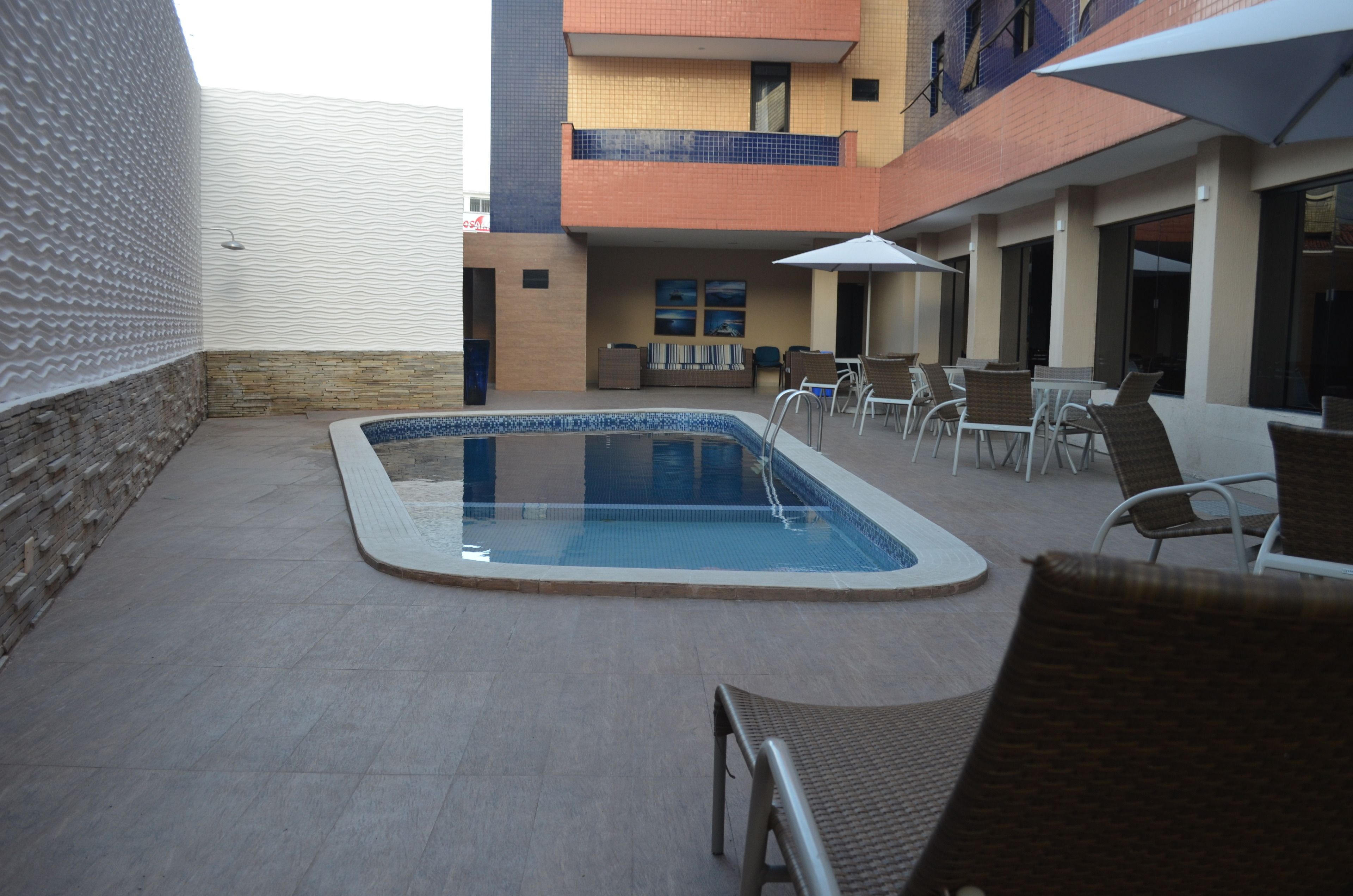 A swimming pool with lounge chairs and umbrellas, surrounded by a brick wall and a building with a tiled balcony.