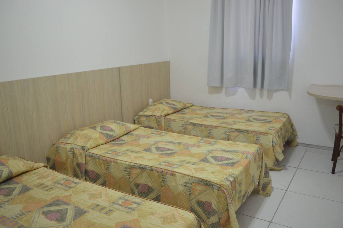 A room with three beds, a wooden headboard, a small table, and a window with curtains.