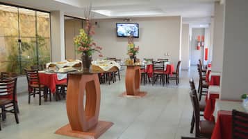 Um restaurante com várias mesas e cadeiras, uma televisão presa à parede e uma parede de pedra com plantas.