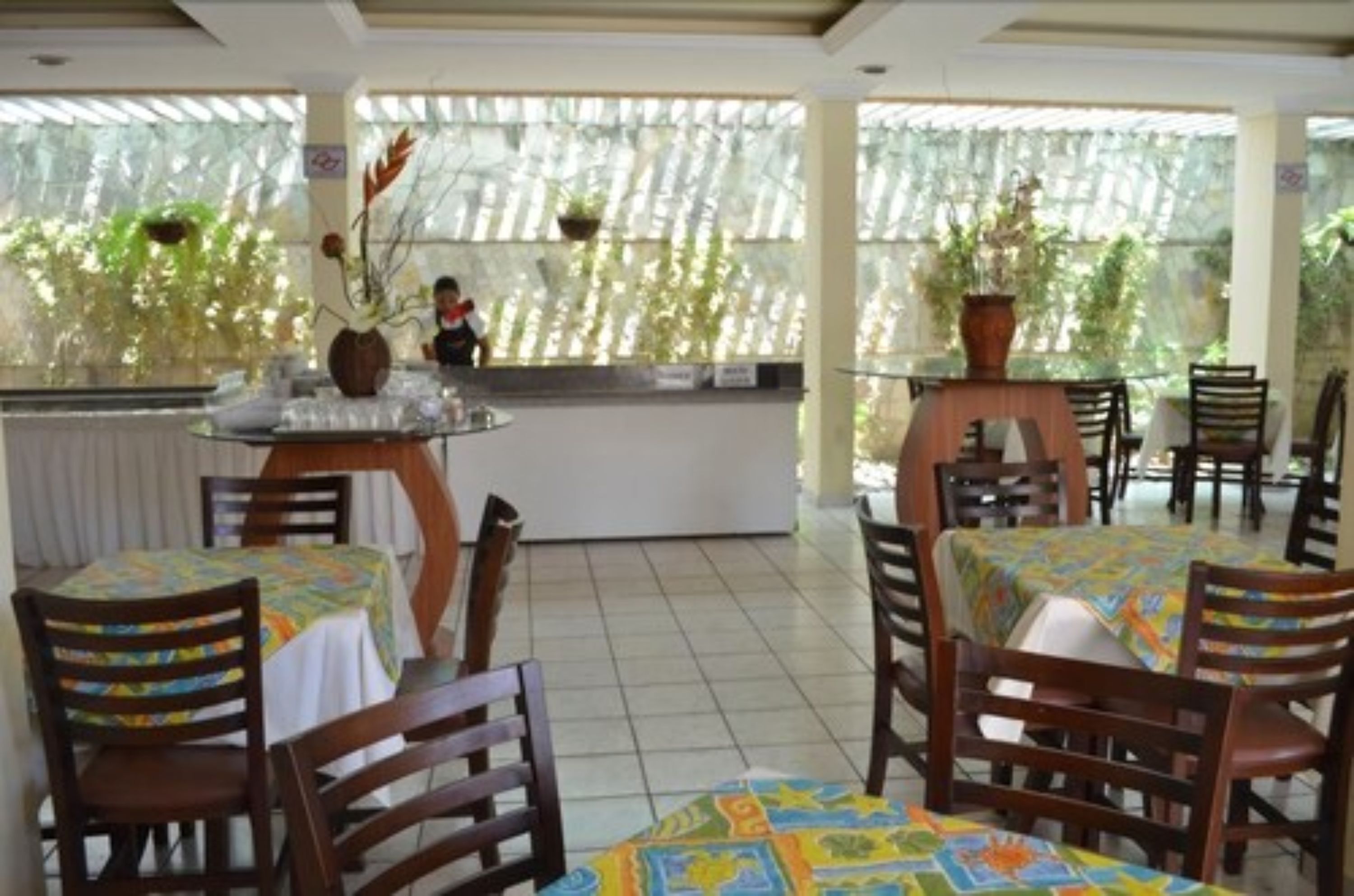 A restaurant with wooden tables and chairs, covered with colorful tablecloths. There are potted plants on the windowsills and a person in the background.
