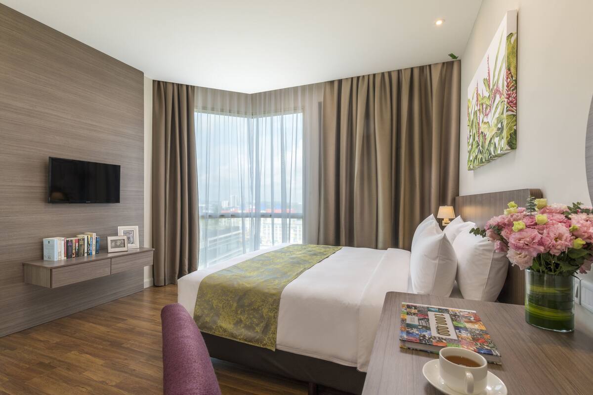 Ein modernes Hotelzimmer mit einem großen Bett, einem flachbildfernseher an der Wand, einem Nachttisch mit einer Zeitschrift und einer Kaffeetasse sowie Blick auf die Stadt durch große Fenster.