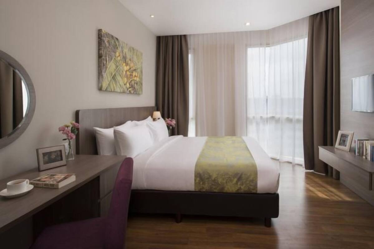 Ein Hotelzimmer mit einem Bett, einem Schreibtisch, einem Sessel, einem großen Fenster mit Vorhängen und einem Bild an der Wand.