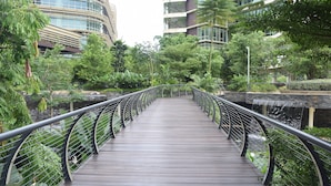 Un puente peatonal de madera con barandillas de metal, rodeado de exuberante vegetación y edificios modernos.