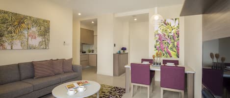 Un salón moderno con sofá, mesa de centro y zona de comedor con sillas. Se observa una cocina al fondo.