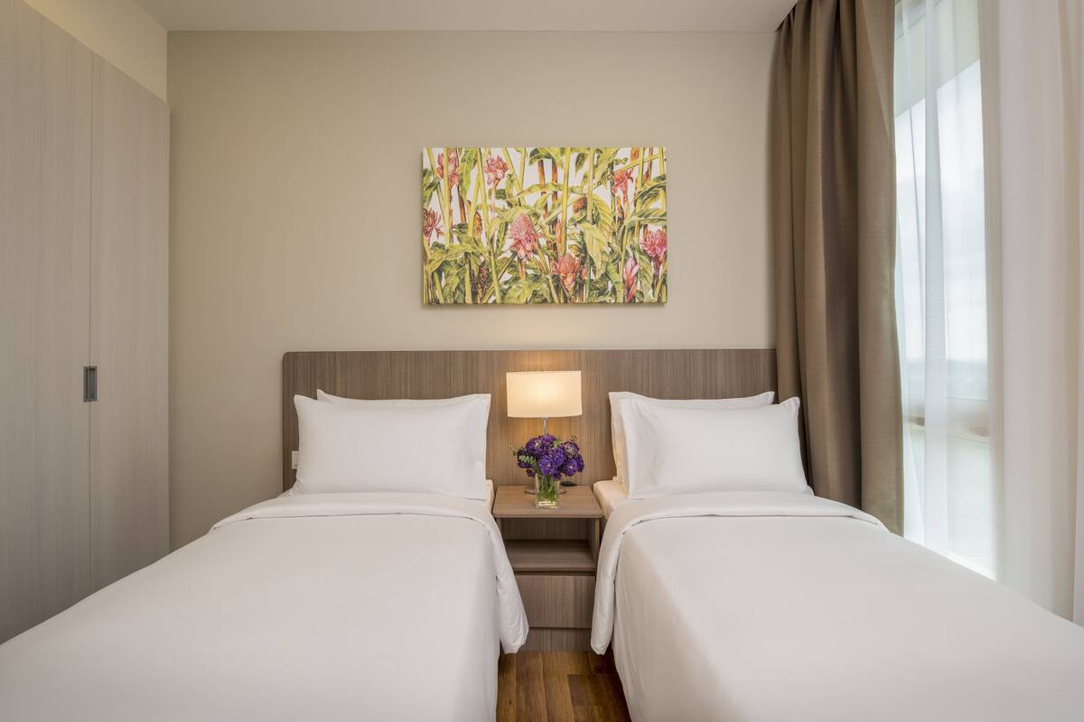Ein Hotelzimmer mit zwei Betten, einem Nachttisch mit Lampe und Blumen, einem großen floralen Kunstwerk an der Wand und einem Fenster mit Vorhängen.