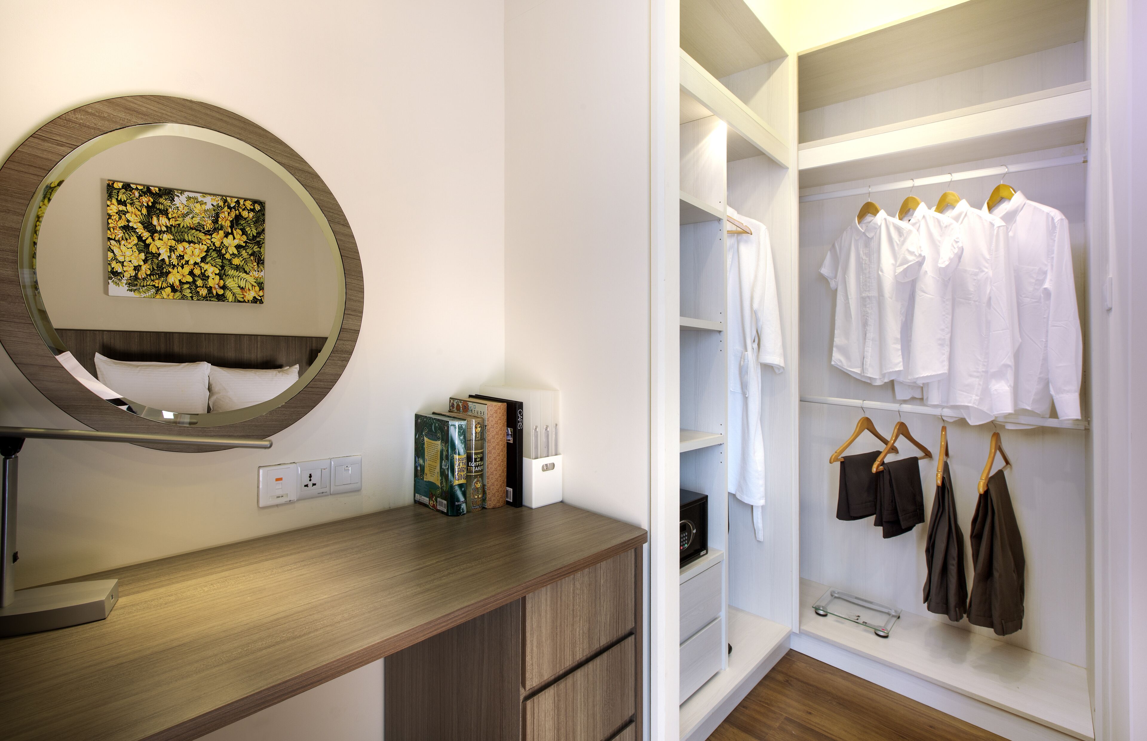A hotel room with a wooden desk, a round mirror, a bed with white linens, a closet with white and brown clothing, and a shelf with books and a small appliance.
