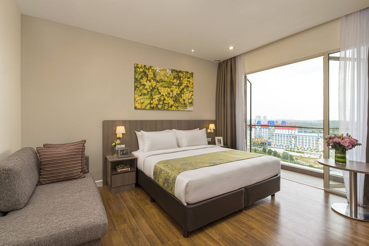 Ein modernes Hotelzimmer mit einem großen Bett, einem grauen Sofa, einem Nachttisch mit Lampen und Blick auf eine Stadtlandschaft durch große Fenster.