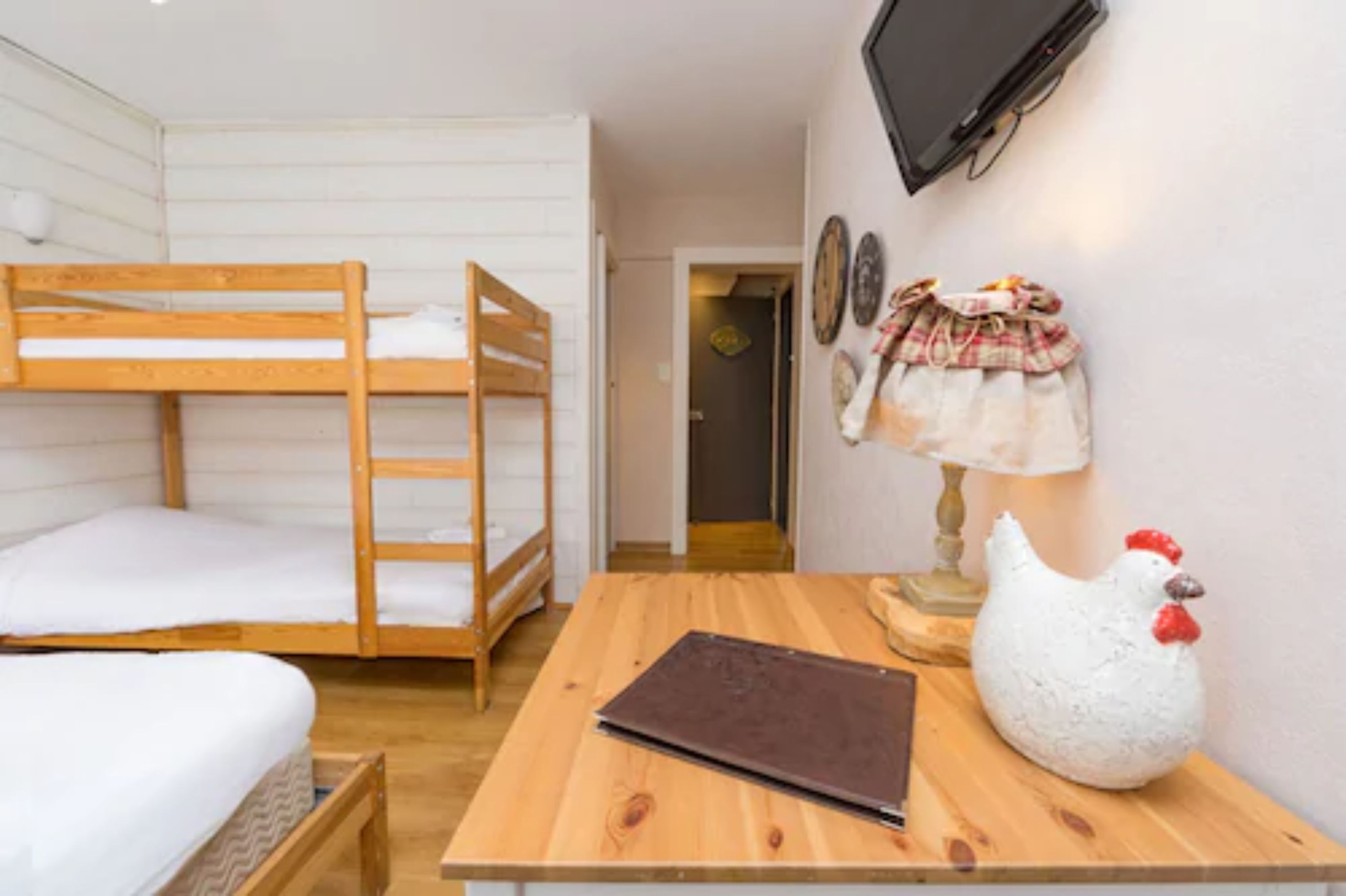 Una habitación con literas, una mesa de madera con una computadora portátil, un adorno de gallina y un televisor montado en la pared.