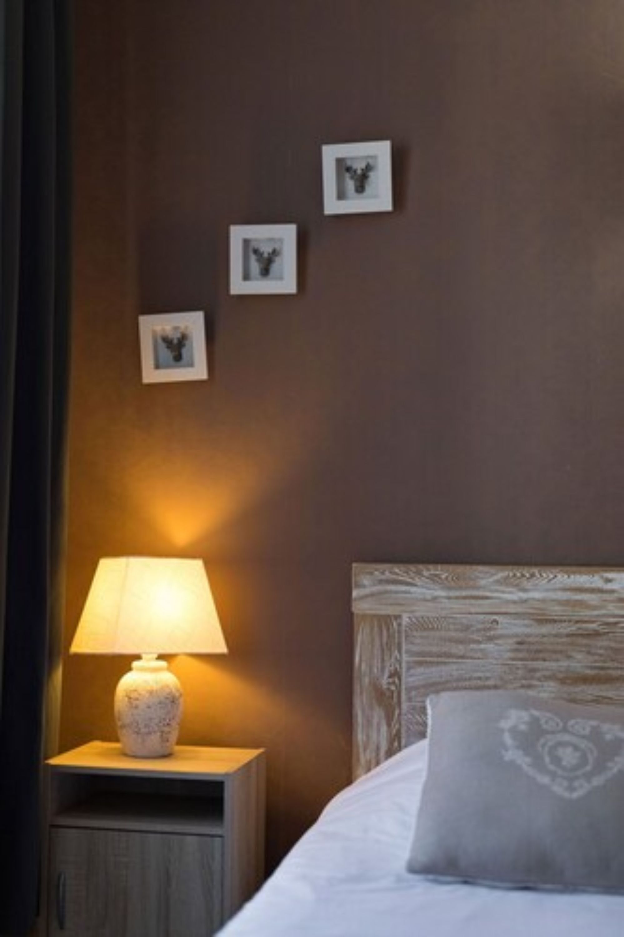 A neatly made bed with a grey pillow, a bedside lamp on a wooden nightstand, and three framed pictures on the wall.