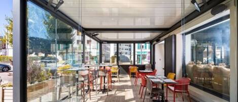 Un área de comedor al aire libre con mesas y sillas, ventanas amplias y vista a la calle.