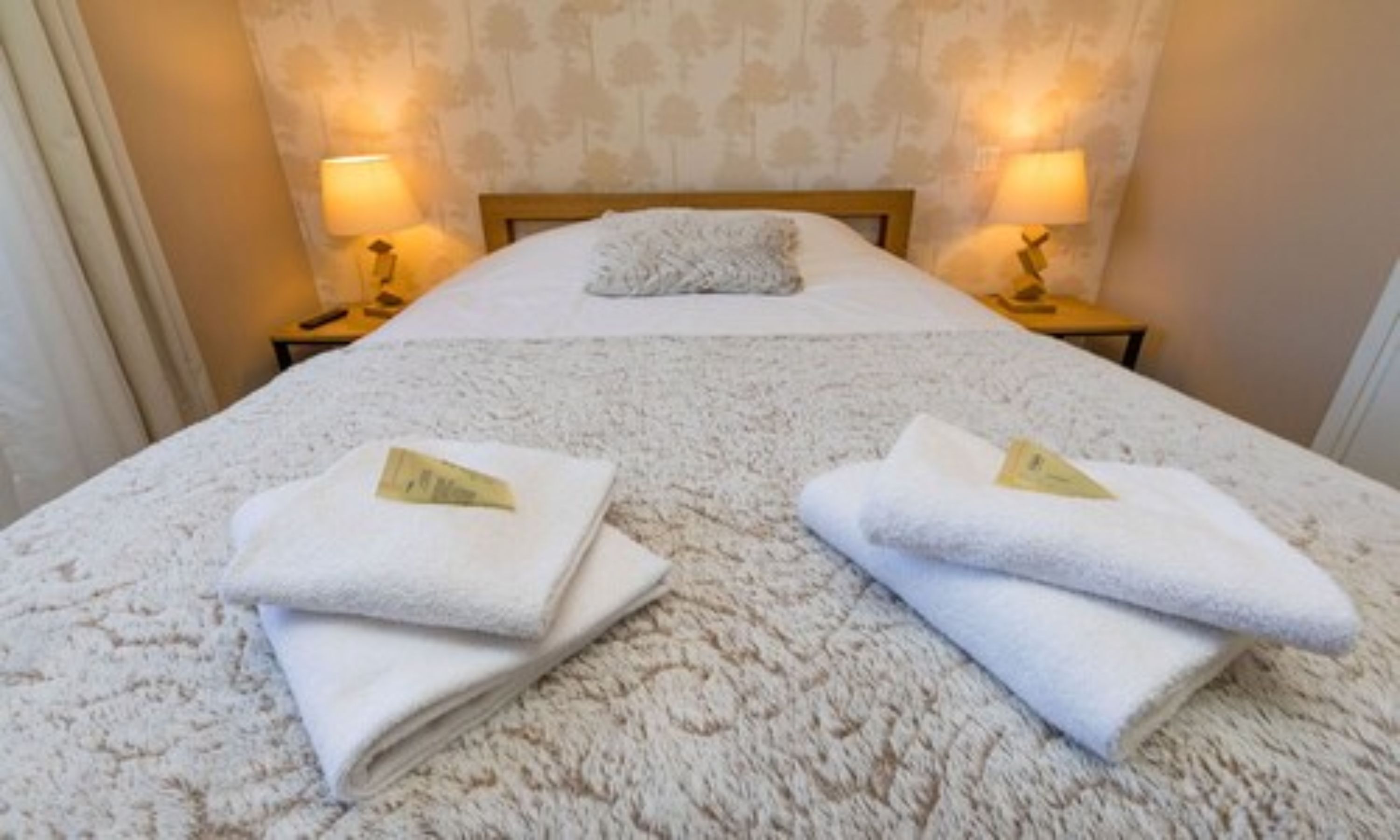A neatly made bed with a white duvet and two folded towels with price tags on a textured carpet.