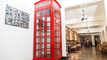 Uma cabine telefônica vermelha em um corredor com uma grande colagem de fotografias na parede e móveis de vime no cômodo.