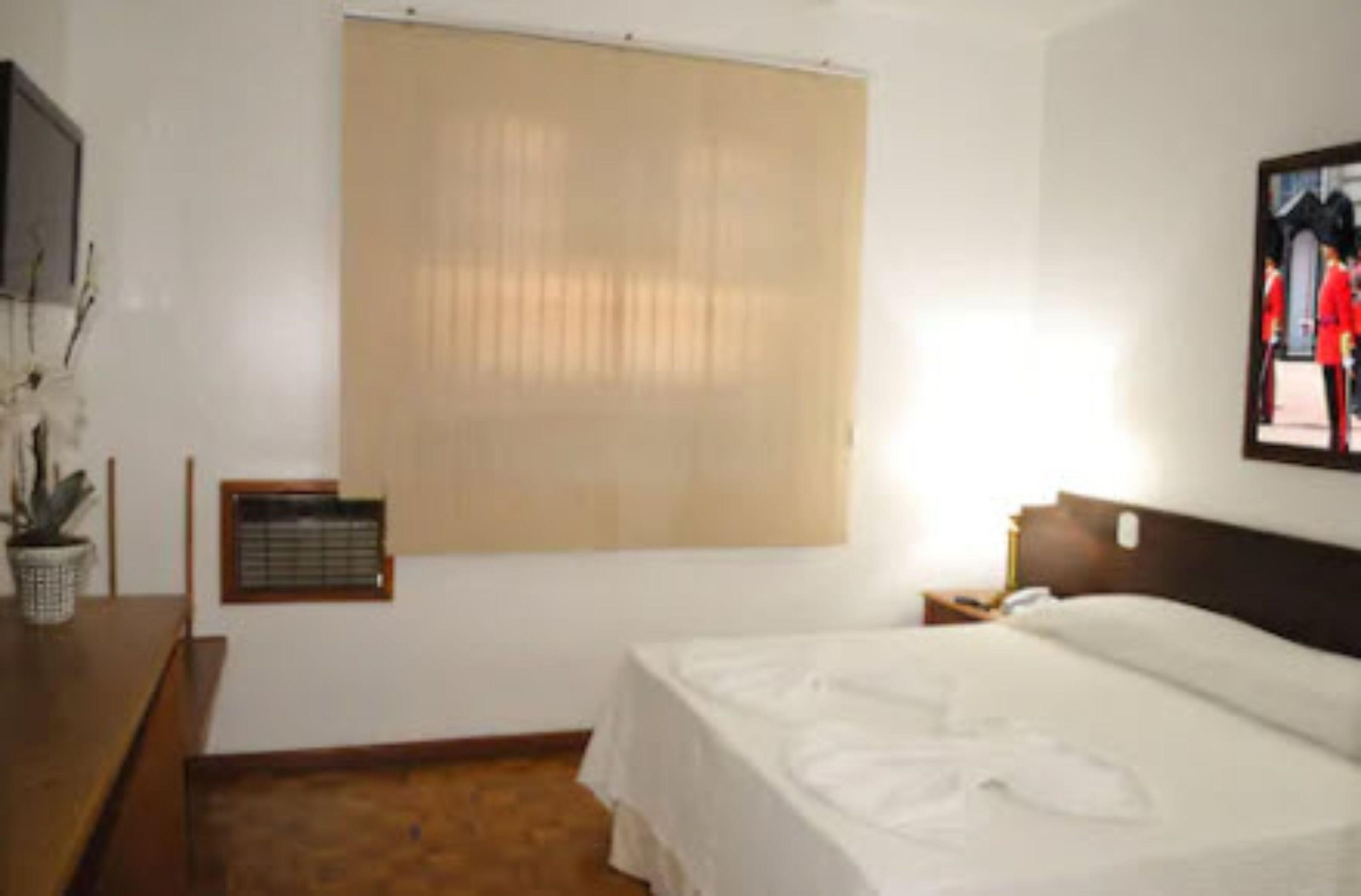 Habitación de hotel con una cama, una ventana con persianas, una mesita con una planta y un cuadro en la pared.