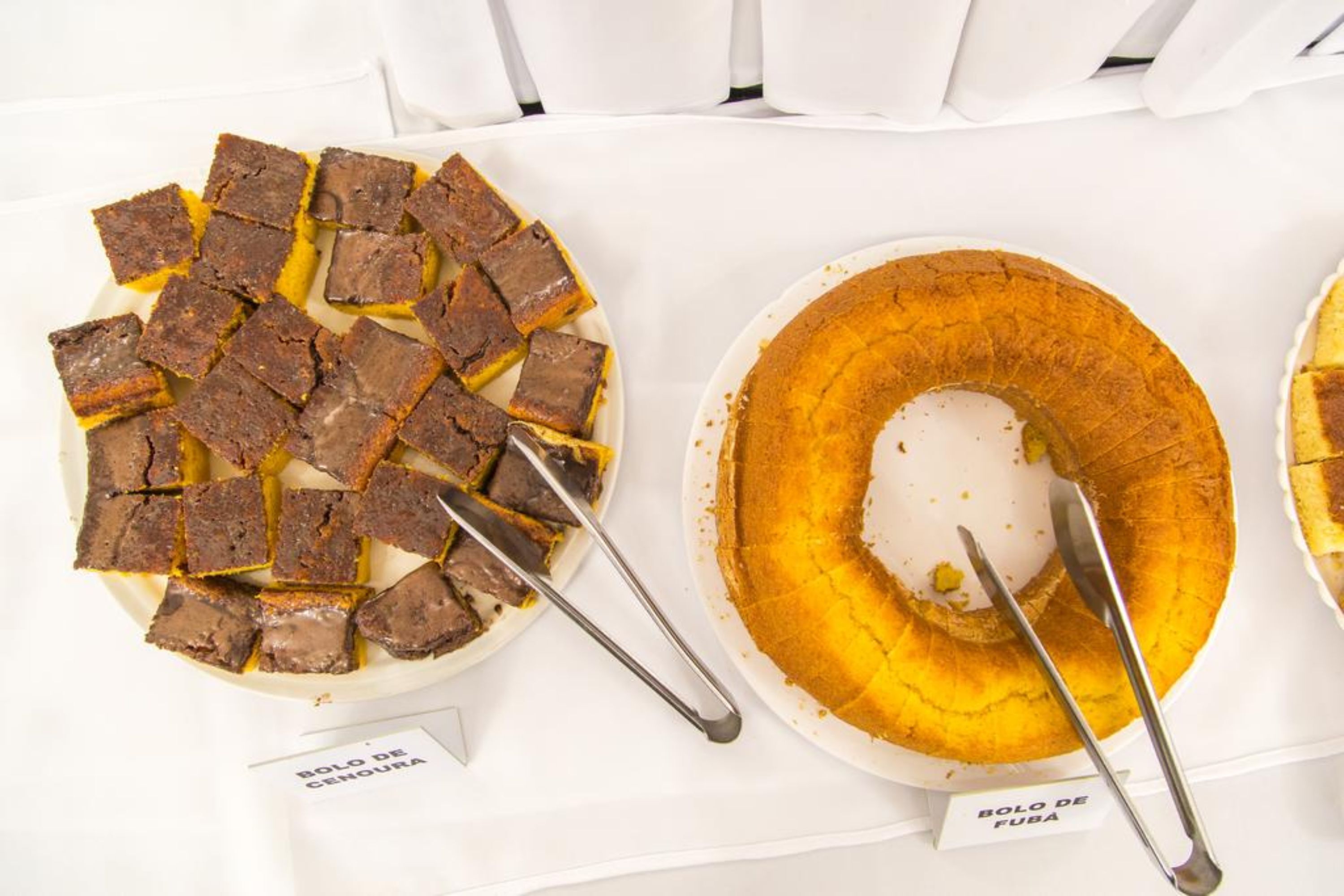 Dos platos de comida, uno identificado con 'BOLO DE CEHURA' y el otro con 'BOLO DE FUBA', sobre un mantel blanco.