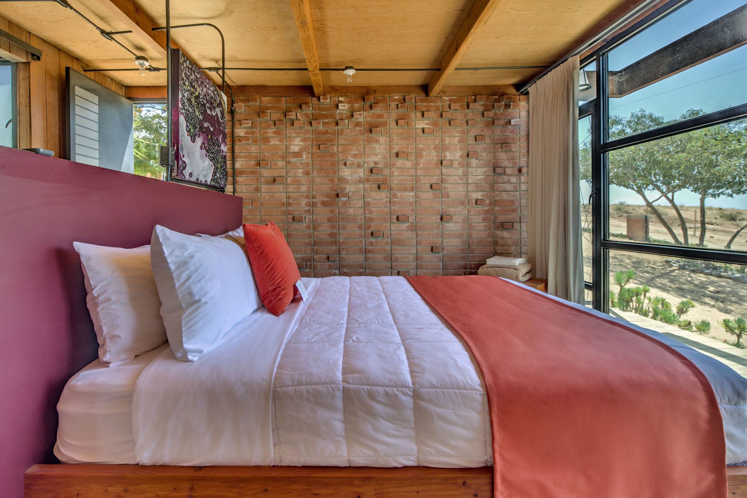 A bedroom with a brick wall, a large window, and a bed with white and red bedding.